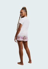 Kortärmad vit t-shirt matchad med rosa shorts som har ett subtilt mönster av enhörningar och volanger i nederkant. Modellen står barfota med en hästsvans.