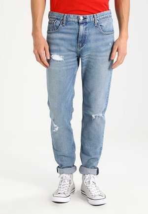 Mężczyzna w jasnoniebieskich podartych dżinsach z podwiniętymi nogawkami i białych wysokich trampkach, stojący na jednolitym tle.