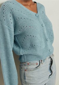 Light blue knitted cardigan featuring a V-neck, button front, and loose fit. The fabric has a textured pattern with small holes for breathability.