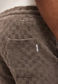 Gros plan sur un short à carreaux marron texturé avec une poche arrière comportant une petite étiquette blanche Lindbergh et une partie d'une chemise blanche au-dessus.