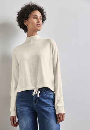 Pull court couleur crème en tissu doux, avec un col montant et un cordon de serrage en bas. Associé à un jean en denim bleu.