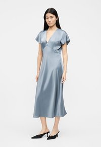 Γυναίκα που φοράει ανοιχτό μπλε σατέν μεσομήκους φόρεμα με κοντά μανίκια και βαθύ V ντεκολτέ, συνδυασμένο με μαύρα mules με μυτερή μύτη και χαμηλό τακούνι kitten heel.