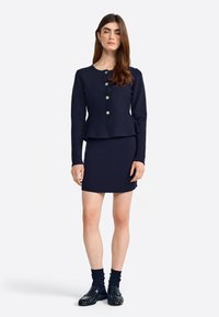 Completo blu navy a due pezzi con giacca peplum aderente caratterizzata da bottoni argentati e gonna a tubino. Indossato con calzini scuri e mocassini.