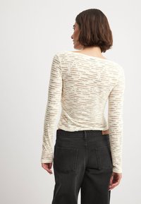 Off-white långärmad topp med en texturerad, utskärningsdesign. Bärs med högmidjade svarta jeans med bakfickor och en läderetikett.