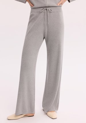 Jasnoszare szerokie dzianinowe spodnie z talią na sznurek, noszone z beżowymi płaskimi butami, osoba stojąca z rękami na biodrach.