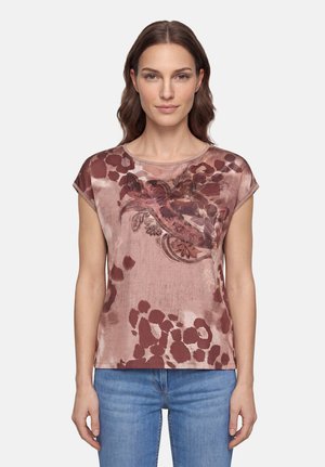 Femme aux cheveux bruns ondulés portant un haut à manches courtes rose et marron à motifs et un jean bleu, face à l'avant sur un fond uni.