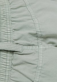 Ljust grönt tyg med en texturerad yta, med samlade rynkor och en sydd söm. Inkluderar en detalj med tygrem.