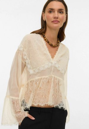 Femme portant un chemisier en dentelle crème translucide avec des manches évasées, un pantalon noir, un collier de perles et des boucles d'oreilles pendantes assorties, les mains dans les poches.