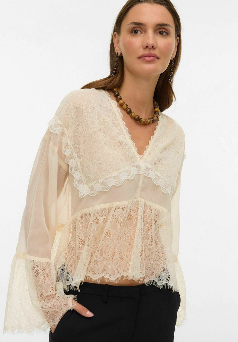 Naine, kes kannab läbipaistvast kreemjas pitsist pluusi lehvatevarrukatega, musti pükse, helmestega kaelakeed ja sobivaid rippuvaid kõrvarõngaid, käed taskus.