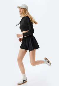 Top negro de manga larga recortado y pantalones cortos de running negros, combinados con zapatillas deportivas blancas y una gorra gris claro, mostrando un diseño elegante.