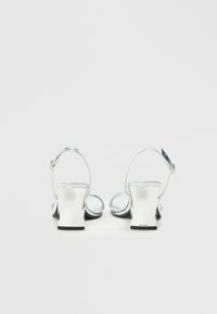 Ασημένιες σανδάλες με φαρδύ τακούνι και καμπύλη ράβδο. Διαθέτουν ρυθμιζόμενο λουράκι αστραγάλου και γυαλιστερή φινίρισμα για επιπλέον λάμψη.