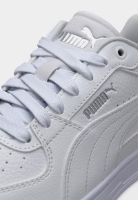 Άσπρο δερμάτινο αθλητικό παπούτσι με υφή στην επιφάνεια, ανοιχτό γκρι κορδόνια, πλευρικό λογότυπο Puma και γυαλιστερές λεπτομέρειες. Κοντινή εικόνα του σχεδιασμού του επάνω μέρους.