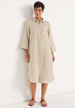 Kvinde med kort mørkt hår iført en beige midikjole med lange ærmer og knapper foran samt brune flade sandaler, stående op ad en hvid væg.