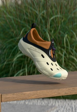 Hellbeige-brauner Crocs-Schuh mit Belüftungslöchern und einer türkisen Zehenklappe, der über einer Holzoberfläche mit grünem Gras im Hintergrund schwebt.