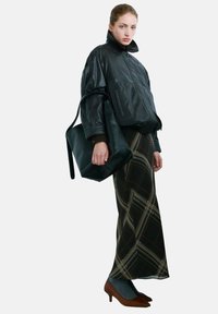 Chaqueta corta de cuero negro con cuello alto, combinada con una falda larga estampada en tonos oscuros y zapatos marrones. Incluye un bolso tote grande negro.