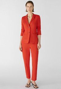 Roter taillierter Blazer mit einem einzelnen Knopf, Reverskragen und Dreiviertelärmeln; passende schlanke Hose. Getragen mit schwarzen Sandalen.
