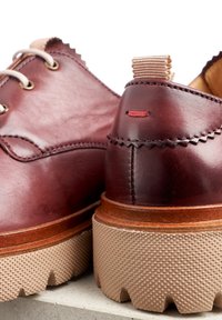 Des chaussures en cuir bordeaux foncé présentent une semelle beige texturée, des coutures décoratives et une languette à l'arrière avec un accent rouge.