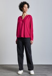 Blusa magenta de manga larga con escote en V y detalle fruncido en los hombros, combinada con pantalones sueltos negros y zapatillas blancas.