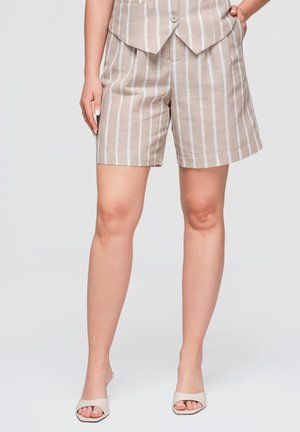 Femme portant un short beige et blanc à rayures verticales avec un gilet assorti et des sandales blanches à bouts ouverts, debout avec une main dans la poche.