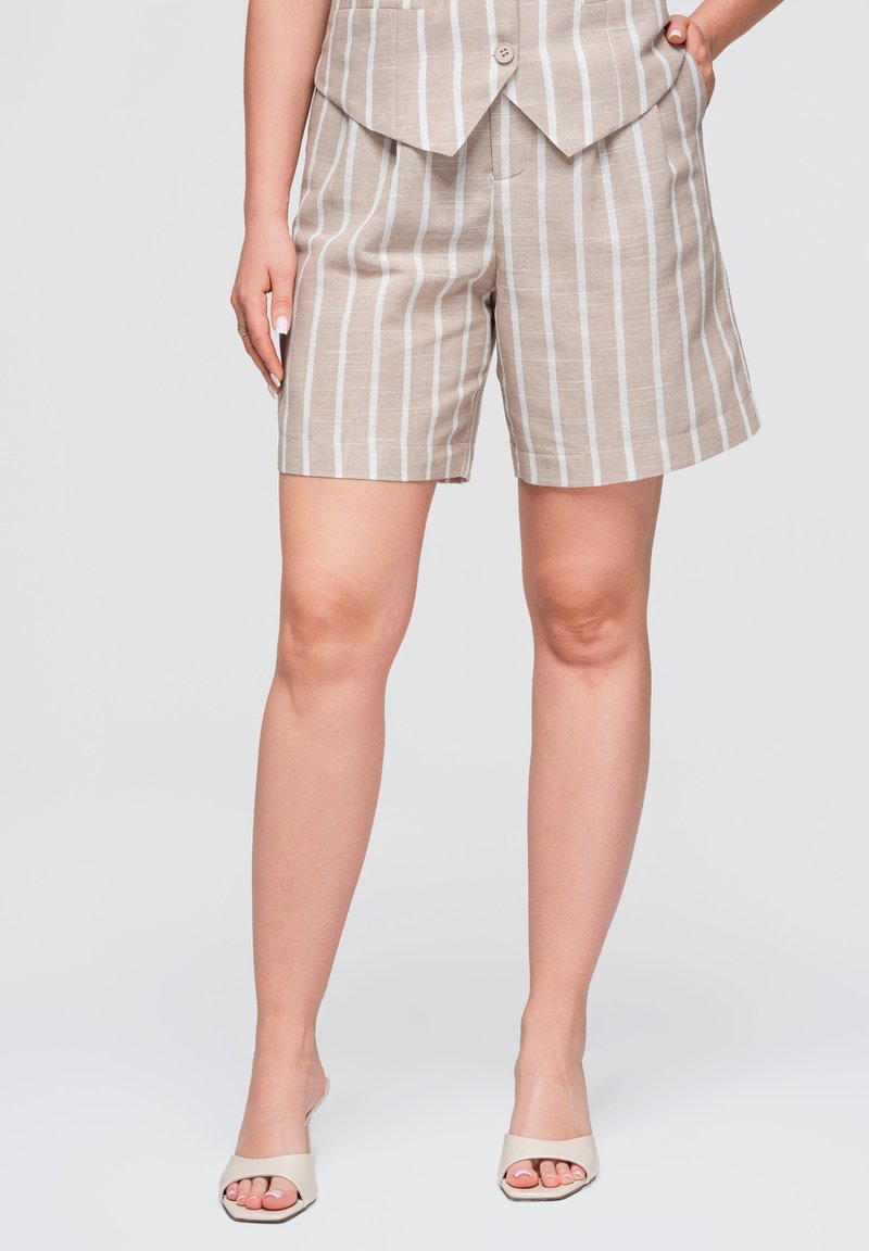 Femme portant un short beige et blanc à rayures verticales avec un gilet assorti et des sandales blanches à bouts ouverts, debout avec une main dans la poche.
