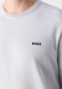 Gros plan d'un pull en maille gris clair avec un col rond et le logo noir "BOSS" sur la poitrine gauche.