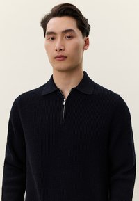 Νεαρός άνδρας με σκουρόχρωμα μαλλιά φοράει μαύρο ριμπ πλεκτό πουλόβερ με γιακά μισής ζώνης, στέκεται μπροστά σε ένα απλό ανοιχτό φόντο.