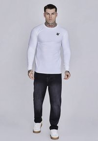 Bela oprijeta majica z dolgimi rokavi in okrolim izrezom ter črnim logotipom, kombinirana s temnimi kavbojkami iz jeansa in belimi supergami z gumijastimi podplati.