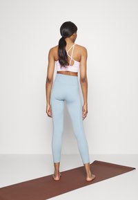 Jasnoróżowy stanik sportowy z krzyżowym plecami w połączeniu z jasnoniebieskimi legginsami typu high-waist. Stoi na brązowej macie do jogi.