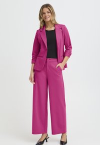 Fukszia öltöny szabott blézerrel és bő szárú nadrággal, sima anyagból. Fekete, testhezálló top alatta. A megjelenést fekete magassarkú cipő egészíti ki.