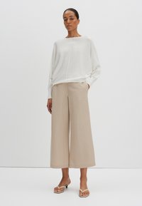 Weißer, locker sitzender Pullover mit weitem Ausschnitt, kombiniert mit beigen Schlaghosen. Sandalen mit minimalistischem Design runden das Outfit ab.
