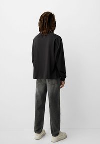 Cămașă lungă, oversized, cu mânecă lungă și croială lejeră, împreună cu blugi șic gri tapered și adidași crem, toate văzute din spate.