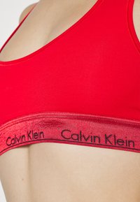 Rød sports-BH laget av glatt stoff, med et bredt elastisk bånd med "Calvin Klein"-logo i svart, enkel design, uten ermer.