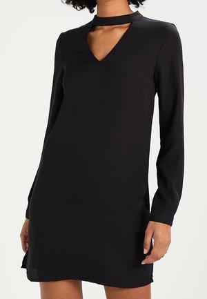 Femme portant une mini-robe noire à manches longues avec un décolleté en V et un col choker découpé.