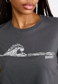 Gray cotton t-shirt featuring a white wave graphic and "ROXY" text, with a crew neck design and smooth texture. Close-up view.