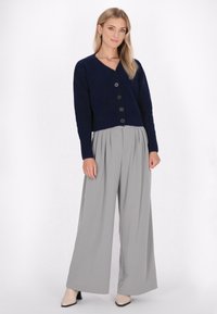 Tummansininen lyhyt cardigan, jossa on napit, yhdistettynä harmaisiin leveälahkeisiin housuihin. Malli seisoo valkoista taustaa vasten beigen värisissä korkokengissä.