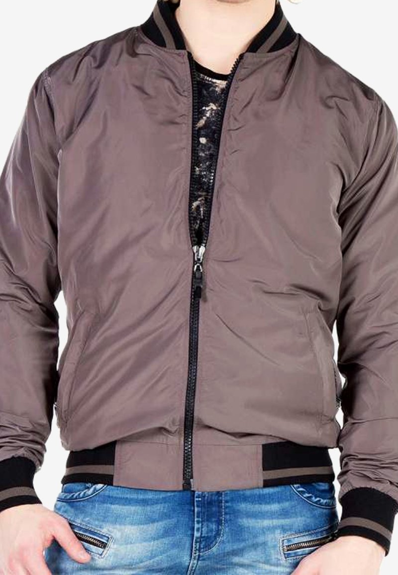 Personne portant une veste bombardier marron avec des poignets et un col noirs, partiellement zippée sur une chemise sombre à motifs, et un jean bleu avec des poches zippées.