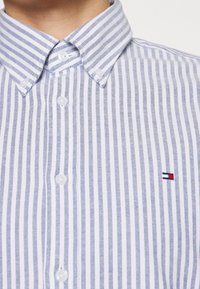 Camicia button-up a righe blu chiaro e bianche, con colletto a punta e un piccolo logo ricamato sul petto. Tessuto testurizzato, design classico.
