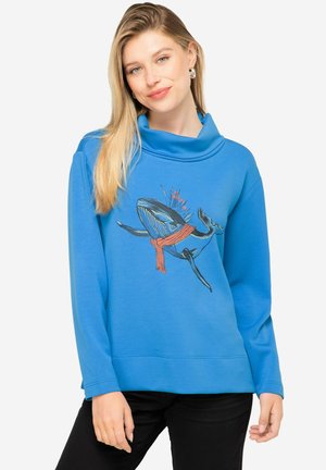 Sudadera azul con cuello en cowl, con gráfico de ballena y una bufanda roja. Textura suave y corte relajado, combinada con pantalones negros.