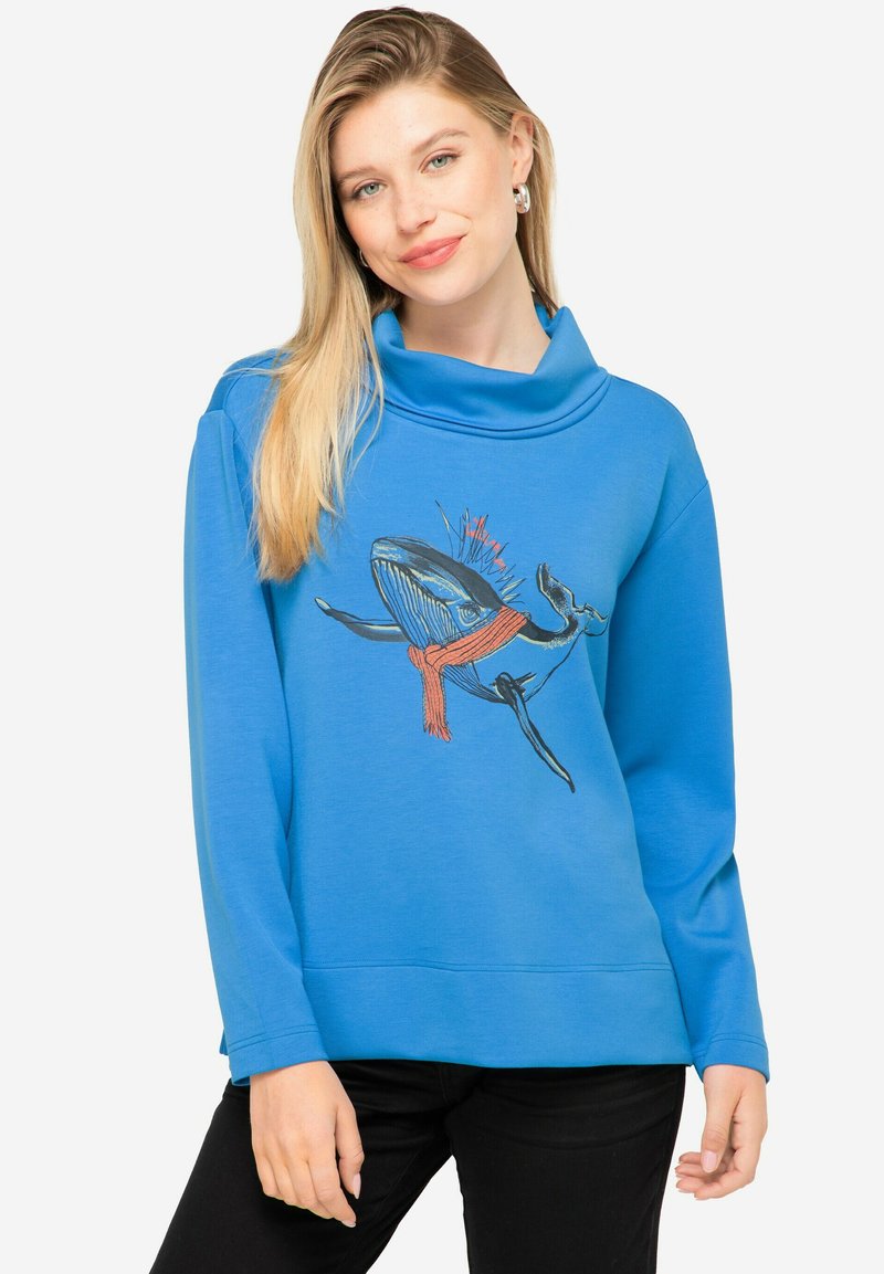 Sudadera azul con cuello en cowl, con gráfico de ballena y una bufanda roja. Textura suave y corte relajado, combinada con pantalones negros.