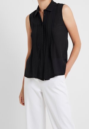 Ärmellose schwarze Bluse mit durchgehender Knopfleiste, plissiertem Design und weicher Textur, kombiniert mit weißen Hosen für einen Kontrast.