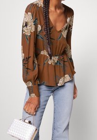 Top peplum marrone con stampa floreale e maniche lunghe, abbinato a jeans azzurri e una borsa bianca trapuntata con hardware dorato.