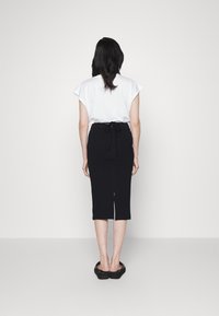 Vit kortärmad topp med breda axlar, kombinerad med en svart figursydd kjol knuten i midjan, som når till halva vaden, och svarta platta skor.