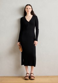 Abito lungo a maniche lunghe in maglina nera a coste con scollatura a V e dettagli con bottoni, caratterizzato da uno spacco laterale. Indossato con sandali intrecciati marroni e una pochette.