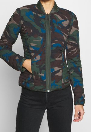 Femme portant une veste matelassée ajustée avec un motif camouflage vert, bleu et marron, fermeture éclair devant, et une main dans la poche.