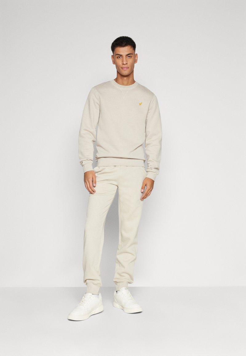 Beige sweatshirt og matchende sweatpants, der har en ribbet rund hals og manchetter, med et lille gult logo på brystet. Sammen med hvide sneakers.