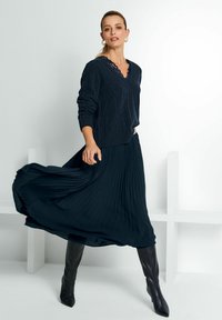 Frau in einem marineblauen Kabelstrickpullover, der mit einem plissierten Rock wirbelt, schwarzen kniehohen Stiefeln und Creolen vor einem weißen Hintergrund.