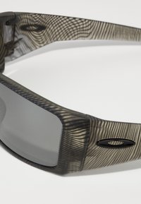 Sonnenbrille mit einem strukturierten grau-schwarzen Rahmen, ausgestattet mit grauen Gläsern und einem schwarzen Logo-Akzent an der Seite.