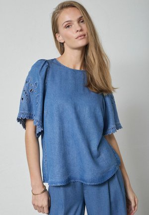 Nuori nainen, jolla on sininen farkkuinen lyhythihainen paita kirjailtuine aukkopeitteineen ja siihen sopivat siniset farkut, seisoo harmaata taustaa vasten.