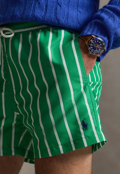 Άτομο που φοράει πράσινη και λευκή ριγέ βερμούδα με μπλε λογότυπο Polo, μπλε πλεκτό πουλόβερ και καφέ δερμάτινο ρολόι στον καρπό.