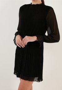 Musta pleatattu mekko, jossa on pitkät puoliläpinäkyvät hihat ja röyhelökaulus. Sisältää joustovyötärön ja levenevän hameen. Suuri silkkinen pinta.